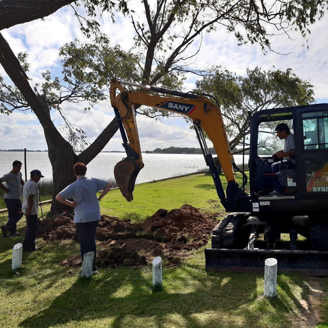 Retroexcavadora abriendo una zanja junto a un lago, con operarios mirando el trabajo